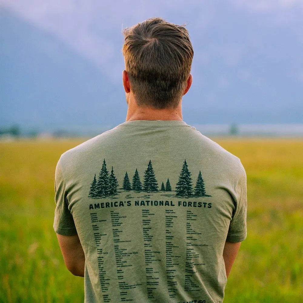 A man viewed from behind wearing an olive green t-shirt with a dark forest graphic and the text &