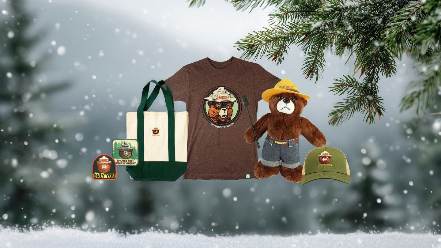 Collection of Smokey Bear-themed merchandise including a t-shirt, bag, plush toy, and cap against a snowy background.