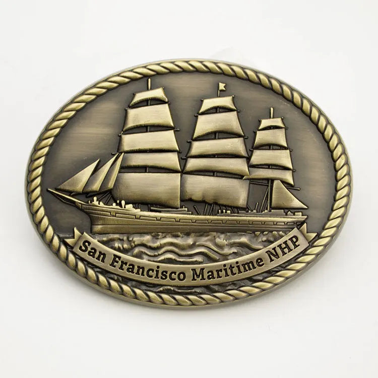A copper colored belt buckle featuring an embossed image of a historic ship with the text 'San Francisco Maritime NHP'.