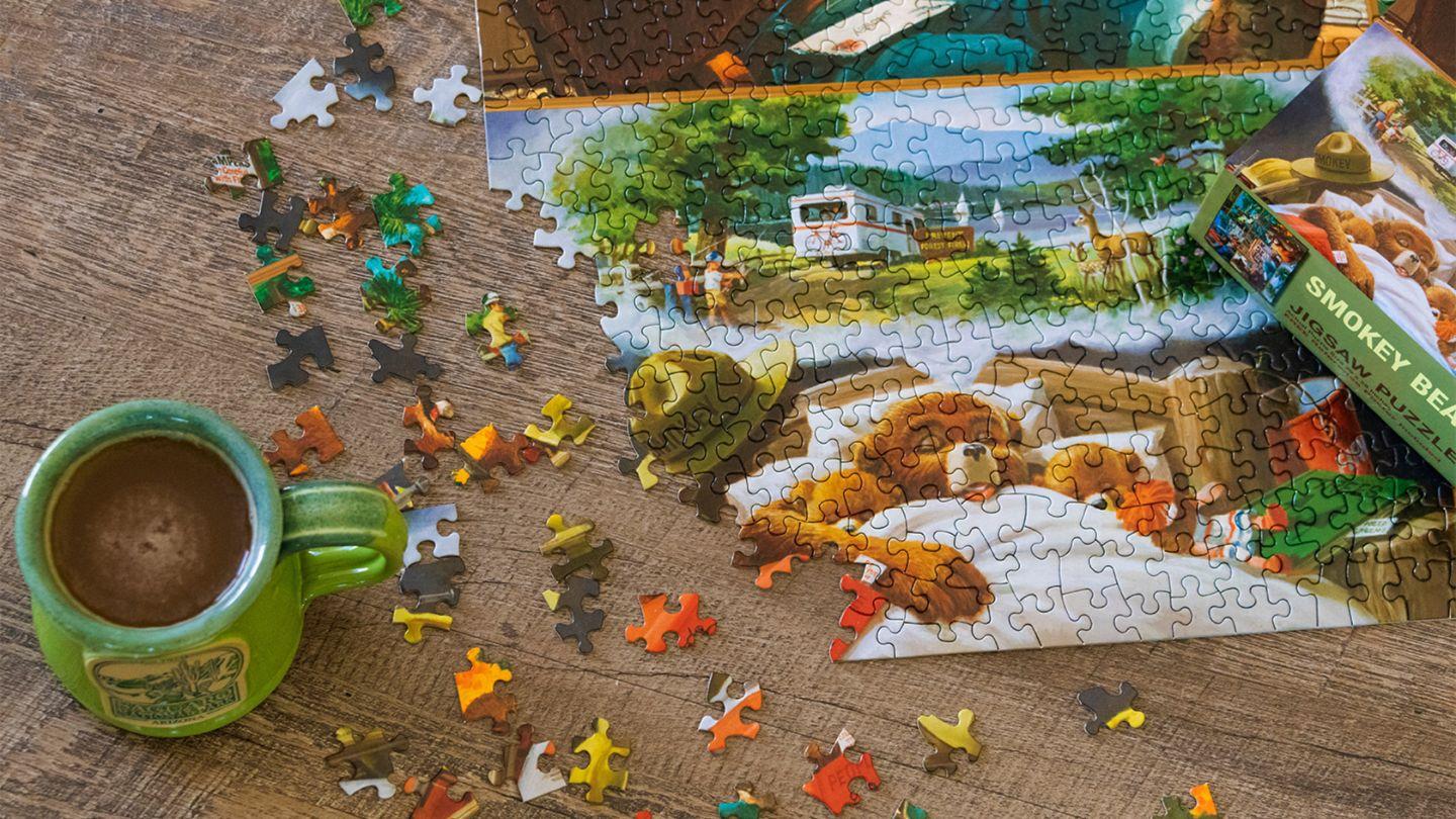 An image of a almost completed Smokey Bear puzzle next to a cup of coffee in a green mug set on a wooden table.