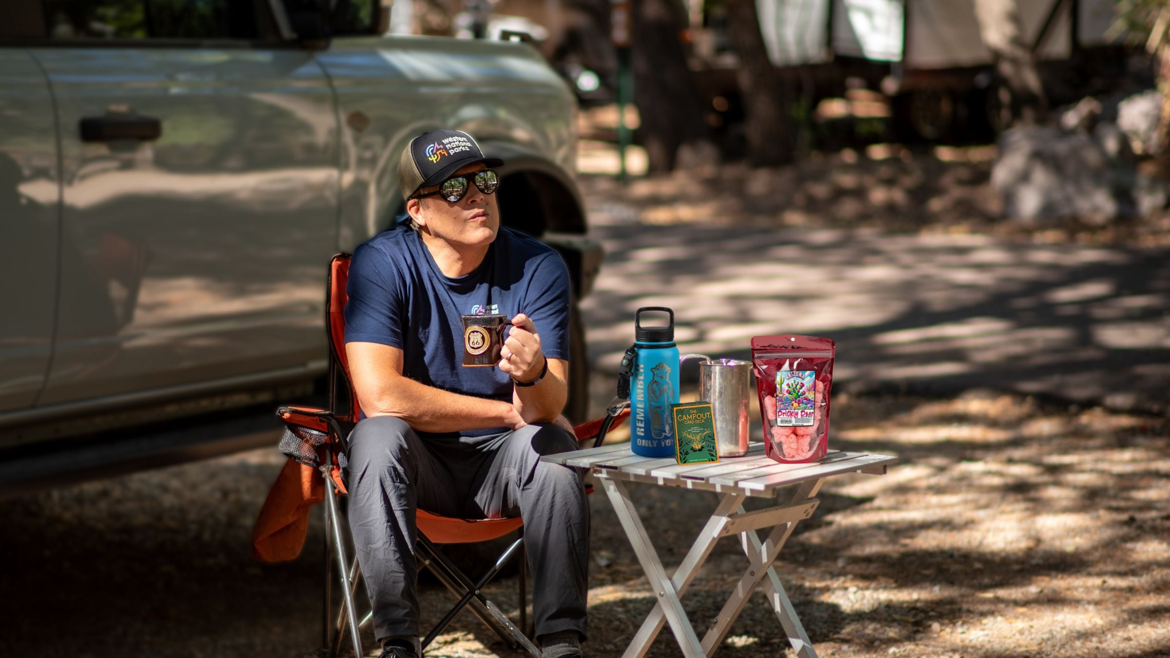 Man sitting at a table outdoors with camping gear, including a water bottle and snacks.